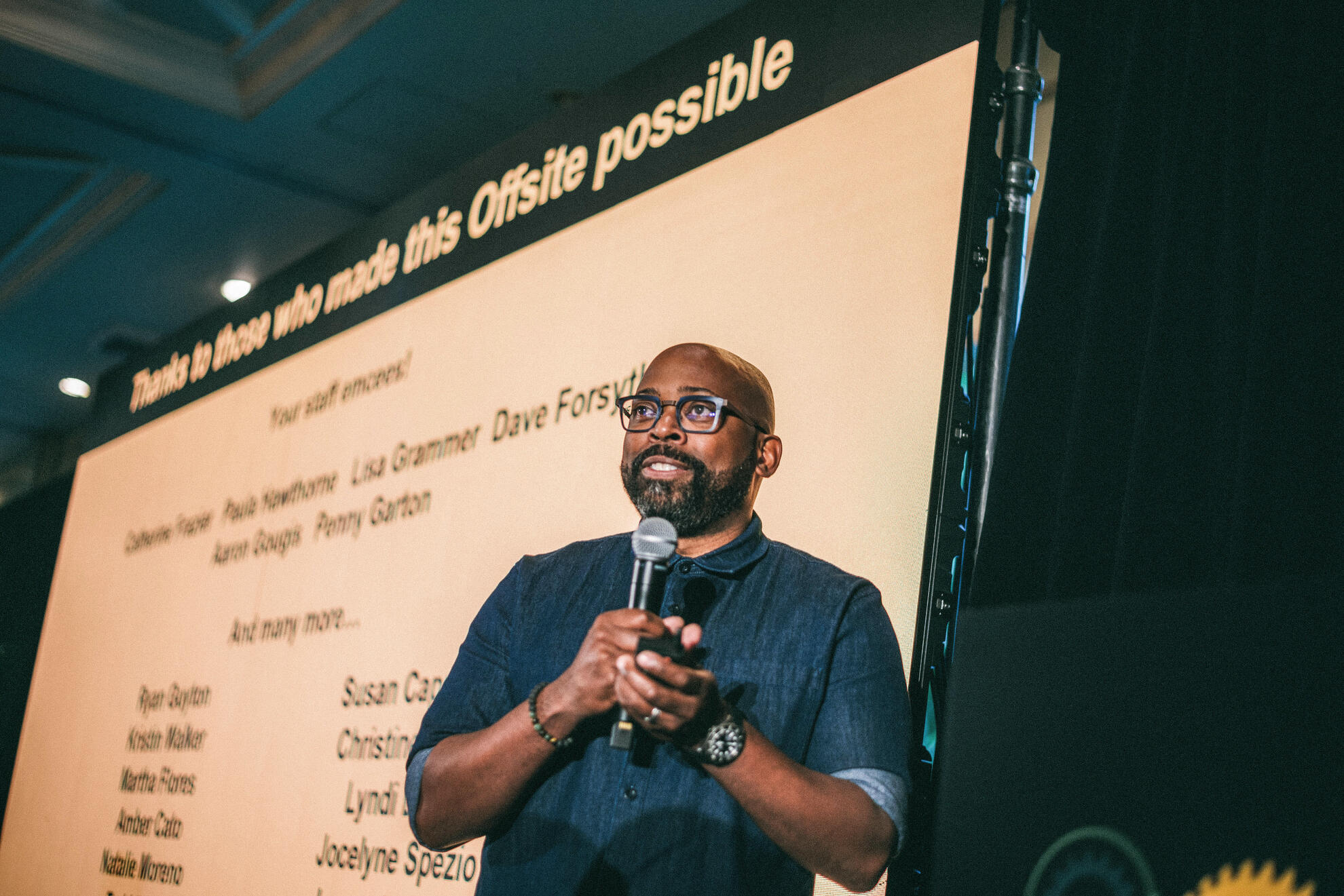 Keynote speaker delivering remarks at a corporate offsite, captured mid-sentence with authentic expression and dynamic stage lighting. The image frames both speaker and presentation slide, emphasizing presence, clarity, and professional tone.